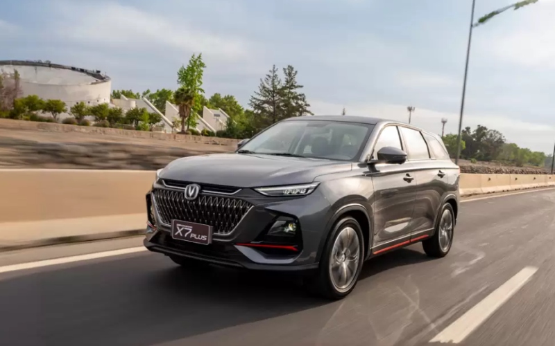 Vista frontal de un Changan X7 Plus gris, un SUV moderno con diseño deportivo, desplazándose por una vía rápida con árboles y un estadio al fondo.
